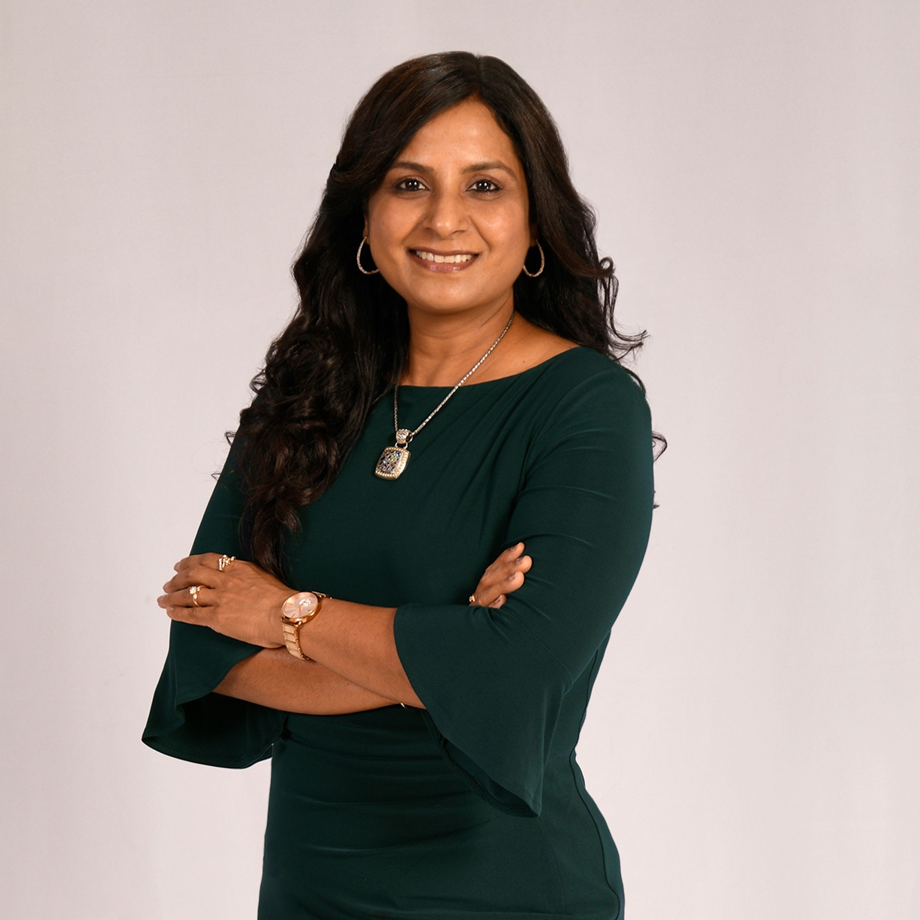 Professional image of a South Asian brown woman, with parted dark brown hair, dark brown eyes, and a smiling friendly face. She is wearing a dark green dress with her arms crossed and her body slightly tilted.